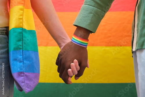 Obraz na płótnie Interracial couple holding hands in front of rainbow flag, celebrating LGBT pride with rainbow wristband w salonie