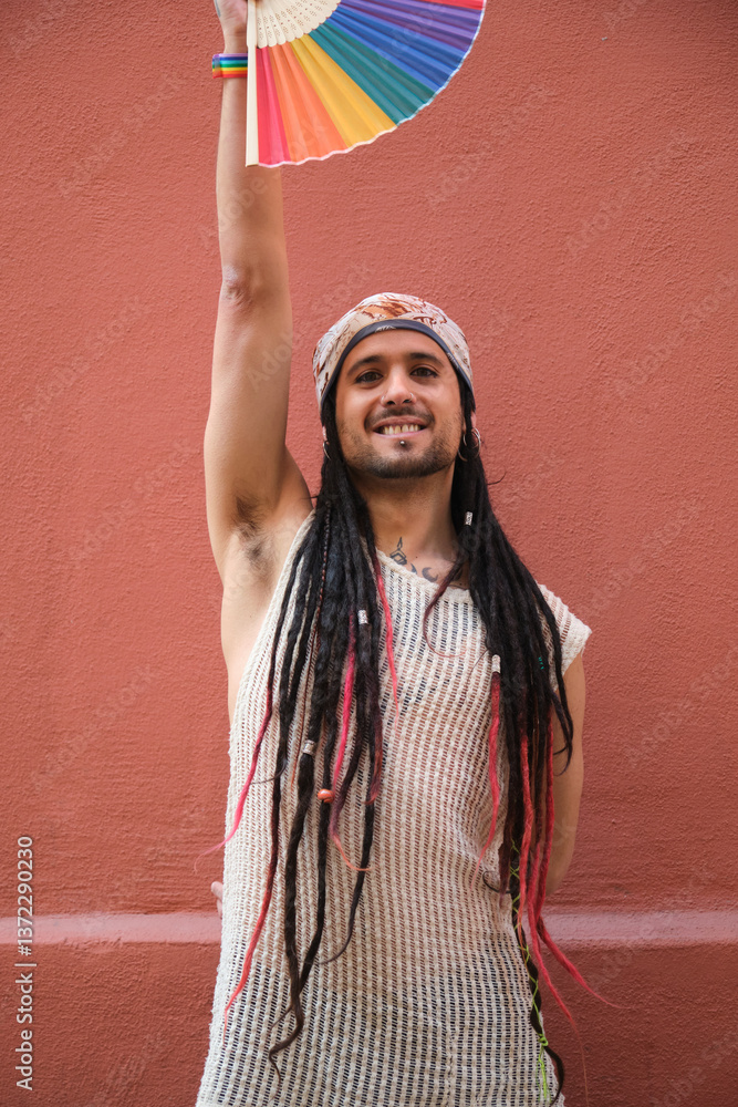 Portrait of smiling young man with dreadlocks and bandana raising rainbow hand fan supporting LGBT community