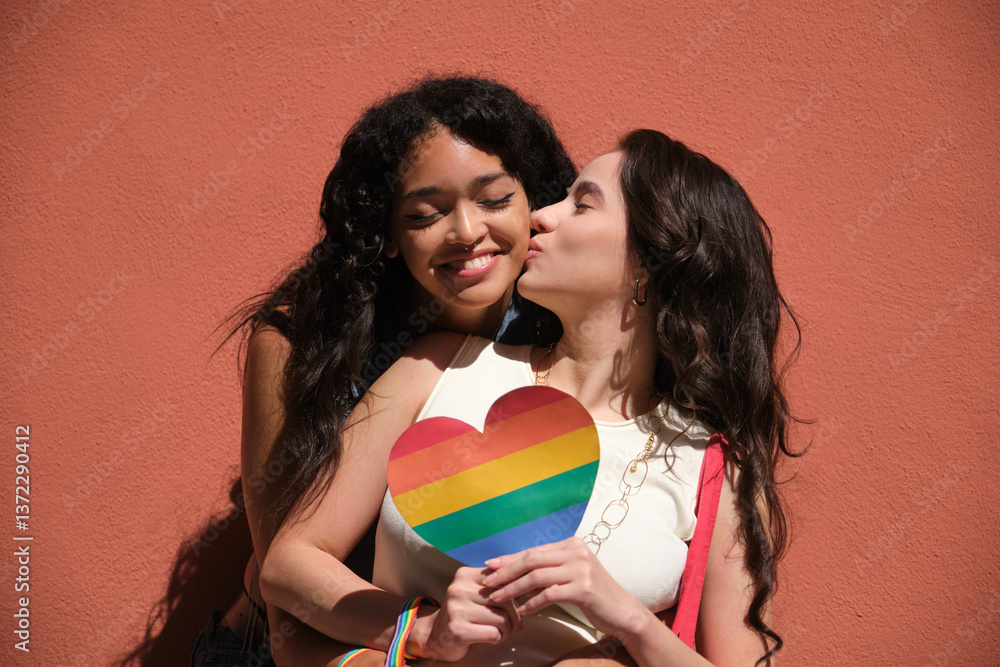 Two young women embracing each other while holding a heart shaped rainbow flag, symbolizing LGBT love and pride