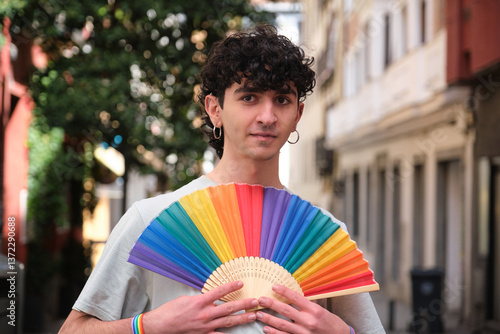 Young curly haired man holding a rainbow fan, celebrating gay pride and supporting LGBT community