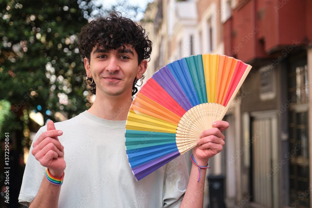 Young man holding rainbow fan and making heart sign with hand wearing a lgbt wristband during pride parade