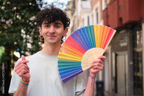 Obraz na płótnie Young man holding rainbow fan and making heart sign with hand wearing a lgbt wristband during pride parade w salonie