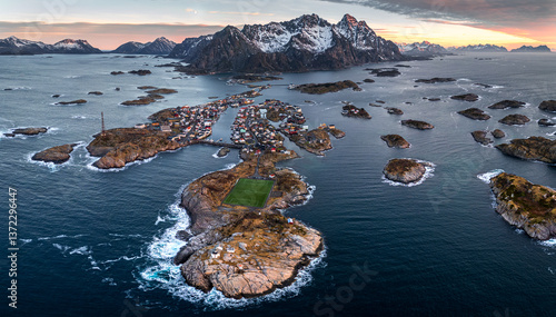Henningsvær auf den Lofoten in Norwegen am frühen Morgen zur goldenen Stunde mit pink-orangefarbenem Himmel, während die Berge im Schnee leuchten und der schneefreie Ort noch im kühlen Schatten liegt