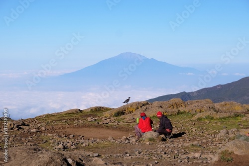 Obraz na płótnie Two men sitting on viewpoint on Shira Camp with Mount Meru in background. Black raven Corvus albicollis (Large-billed raven) on rocks. Machame Route, Tanzania w salonie