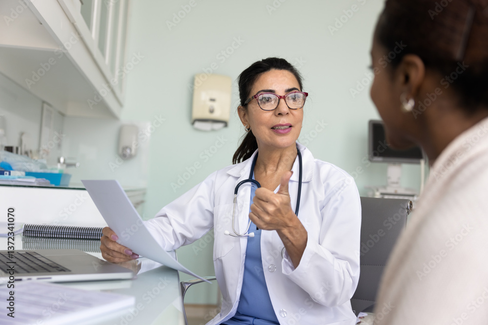 Friendly engaged middle aged elder doctor giving consultation to African patient, talking to young woman in office, holding paper medical report, history, giving prescription, explaining treatment Friendly engaged middle aged elder doctor giving consultation to African patient, talking to young woman in office, holding paper medical report, history, giving prescription, explaining treatment