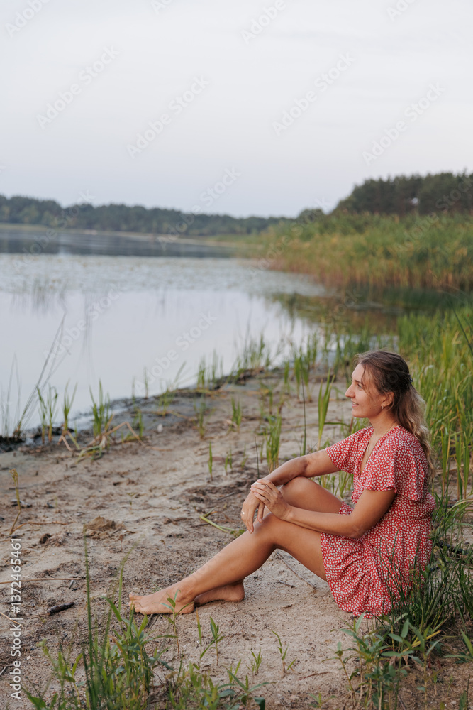 Fototapeta premium A woman in a red dress sits barefoot on the lakeshore, surrounded by wild nature. She smiles and enjoys the tranquility, creating an atmosphere of harmony, relaxation, and connection with nature