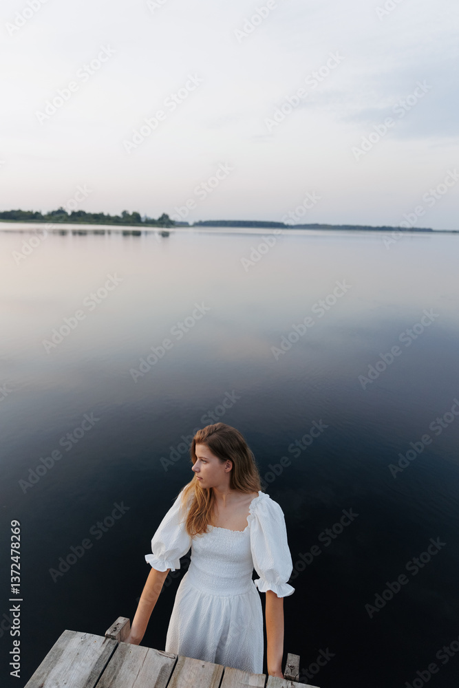 Fototapeta premium A woman in a white dress stands in the water near a wooden pier, holding onto the edge. The evening sky reflects in the calm lake. In the distance, a boat with people adds to the sense of solitude