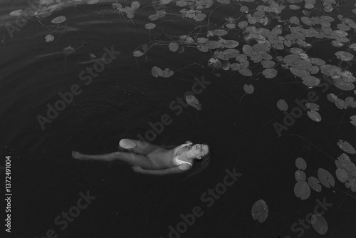 A black-and-white photo of a woman in a swimsuit floating peacefully on her back among water lilies in dark water. Her face reflects harmony and serenity, creating an atmosphere of calm and mystery.