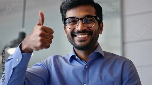 A confident man wearing glasses and a blue shirt gives a thumbs-up gesture, radiating positivity and enthusiasm. The professional setting suggests success or satisfaction in a work-related context.