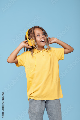 Trendy little boy with african dreads listening music in headphones dancing over blue background