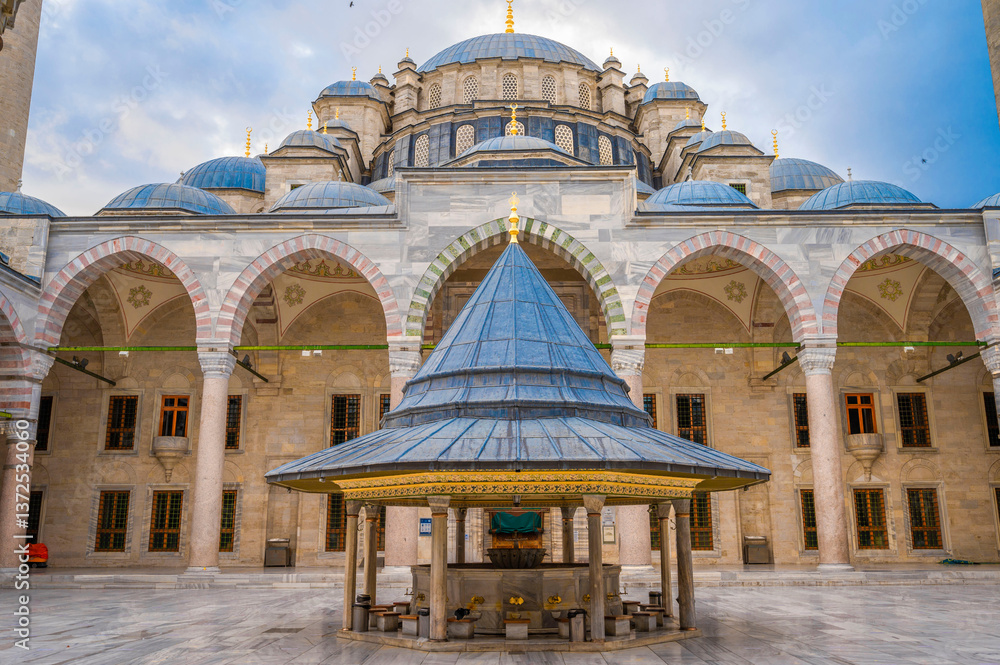 Fototapeta premium Majestic view of the inner courtyard and ablution fountain of the Nuruosmaniye Mosque in Istanbul showcasing Ottoman architecture with domes, arches, and intricate designs under a cloudy blue sky