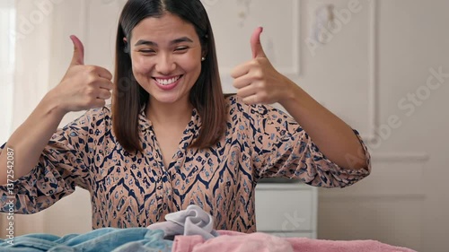 Cheerful woman in shirt shows thumbs up. Cozy environment suggests that she is sorting out newborn baby clothes, radiating joy and excitement.