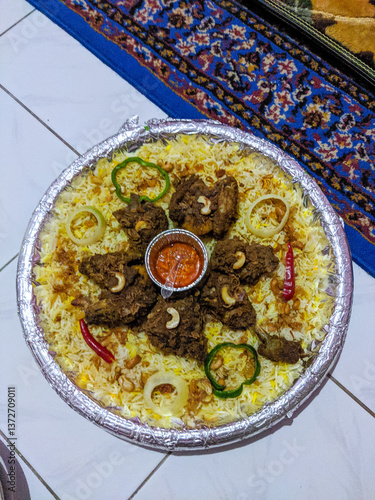 Platter of Middle Eastern Kabsa or Mandi with fragrant rice, tender meat, nuts, sliced vegetables, and a bowl of sauce, served on a patterned surface, showcasing rich flavors and tradition