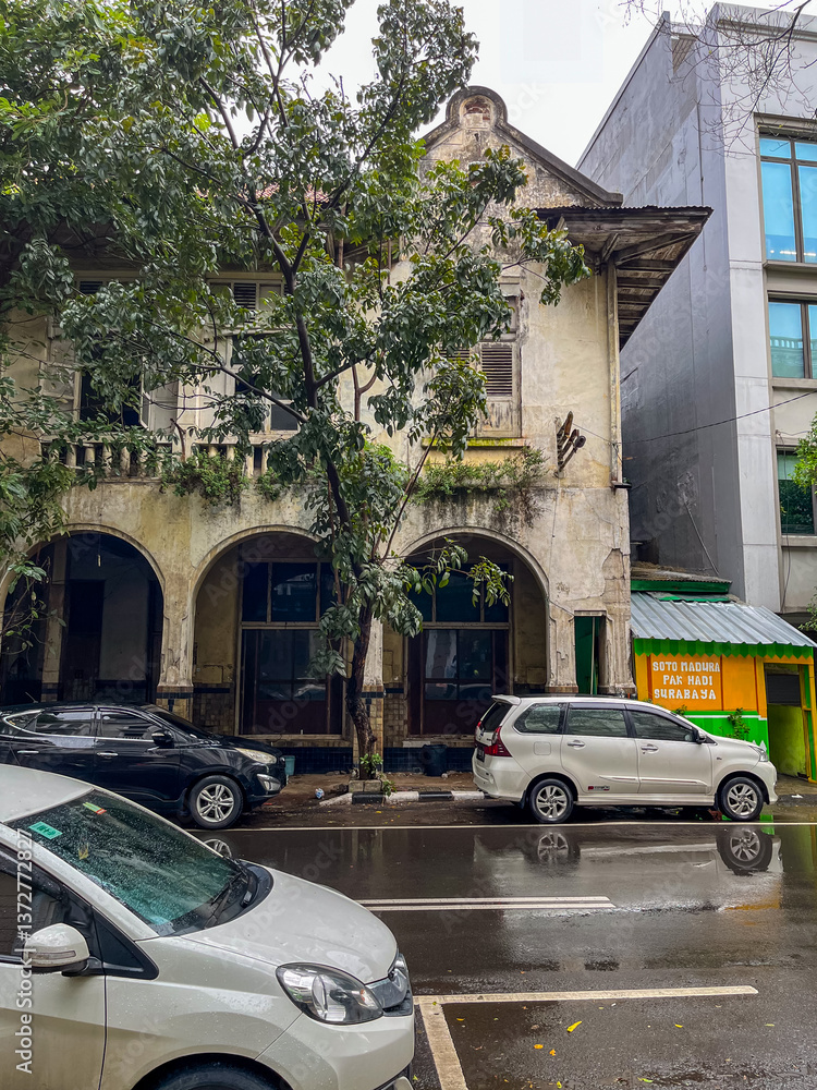 Old weathered two-story building with arched openings stands beside a modern structure on a wet Jakarta street, blending historic charm with urban life and parked cars.