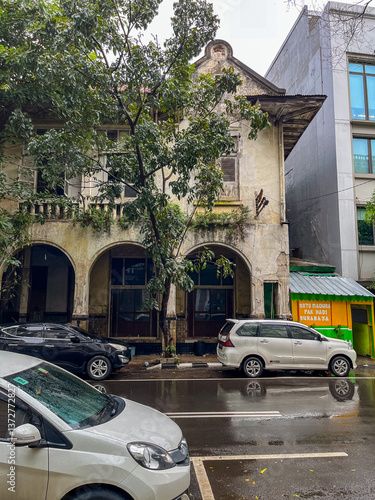 Obraz na płótnie Old weathered two-story building with arched openings stands beside a modern structure on a wet Jakarta street, blending historic charm with urban life and parked cars. w salonie