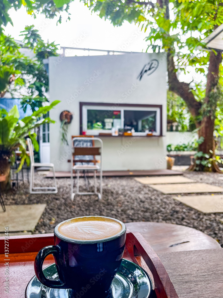 Outdoor café features a small white building with a service window, surrounded by lush trees and greenery. Stone pavers lead to cozy seating areas, creating a relaxed and natural atmosphere.