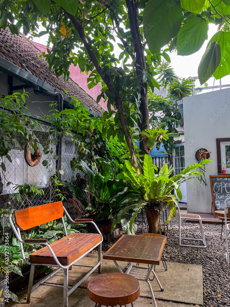 Outdoor café features a small white building with a service window, surrounded by lush trees and greenery. Stone pavers lead to cozy seating areas, creating a relaxed and natural atmosphere.