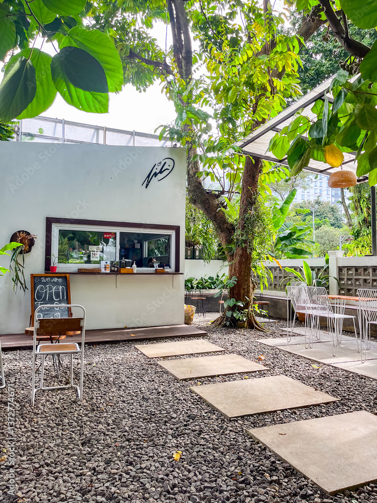 Outdoor café features a small white building with a service window, surrounded by lush trees and greenery. Stone pavers lead to cozy seating areas, creating a relaxed and natural atmosphere.