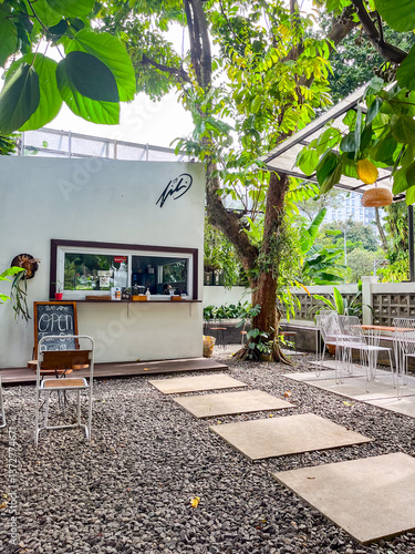 Outdoor café features a small white building with a service window, surrounded by lush trees and greenery. Stone pavers lead to cozy seating areas, creating a relaxed and natural atmosphere.