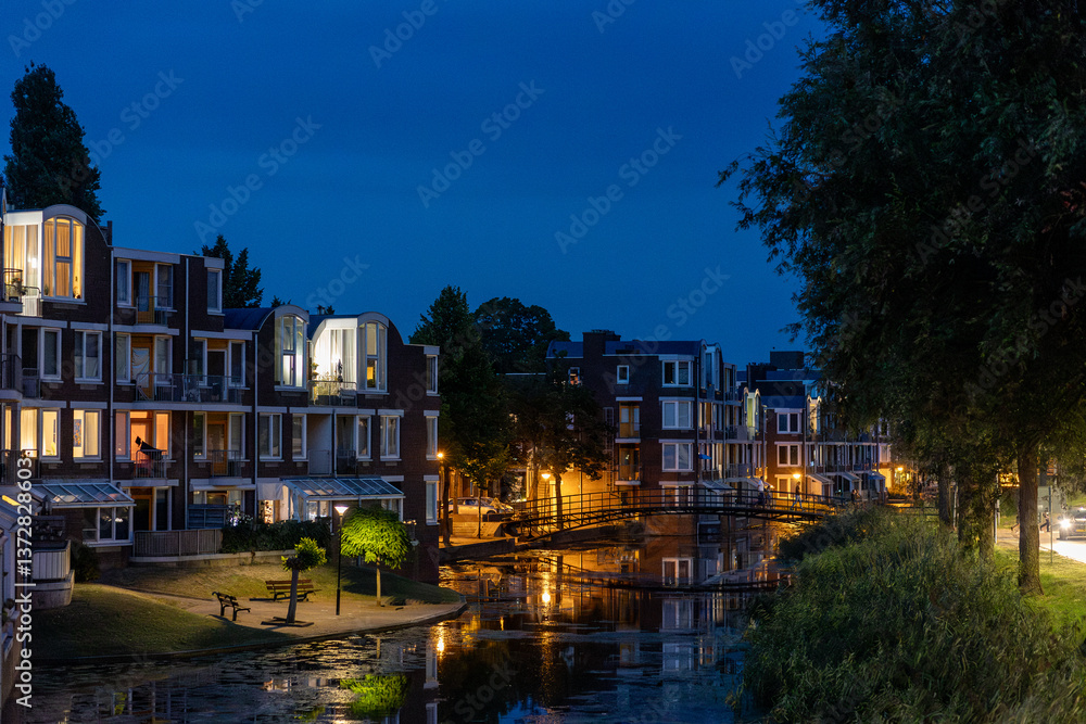 Fototapeta premium A serene canal scene at dusk, with modern brick buildings illuminated by warm lights. Reflections shimmer on the water, while trees and a narrow walkway add natural charm to the urban setting.