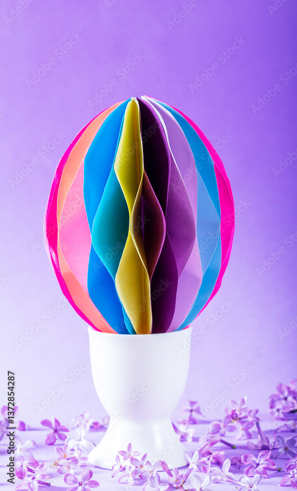 Decorative paper Easter egg in an egg cup and lilac flowers on a lilac background.