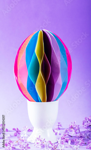 Decorative paper Easter egg in an egg cup and lilac flowers on a lilac background.