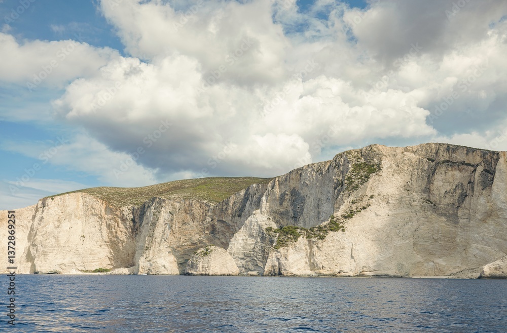 Naklejka premium Coastal cliffs rise majestically above calm waters under a partly cloudy sky, embodying the beauty of summer in Greece. The tranquil scene invites relaxation and exploration