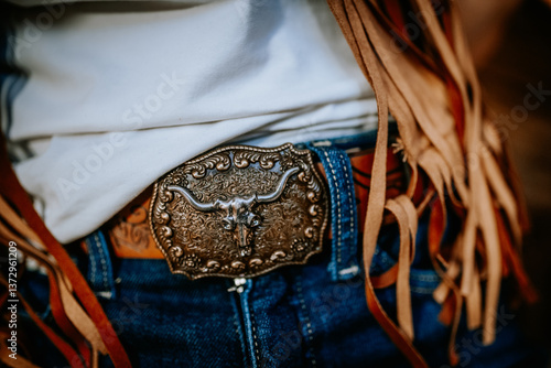 Photography Close-up of an ornate Western-style belt buckle featuring a longhorn skull, worn with denim jeans and fringed suede vest, evoking cowboy and rodeo aesthetics