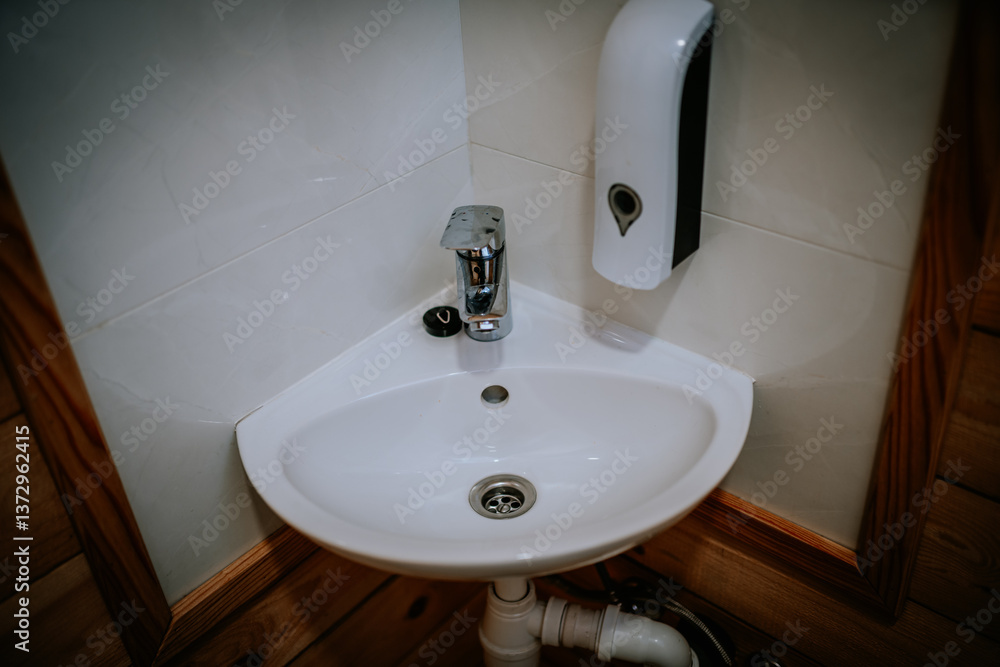 Fototapeta premium Corner bathroom sink with chrome faucet, soap dispenser, and exposed plumbing, set against tiled and wood-paneled walls in a compact washroom setup..