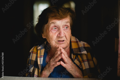 A portrait of an elderly woman sitting at a table at home, her hands clasped and emotions reflecting in her thoughtful expression.