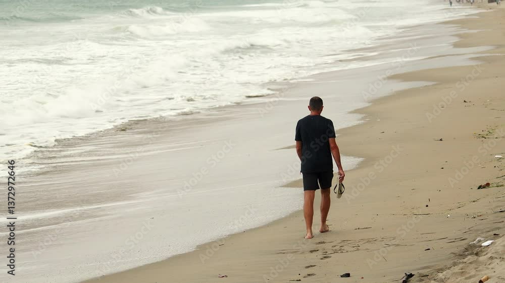 Back view of unrecognizable man in black T-shirt and shirts walks along beach with storms and big waves. walk to refresh his thoughts and merge with nature. Back view of unrecognizable man in black T-shirt and shirts walks along beach with storms and big waves. walk to refresh his thoughts and merge with nature.