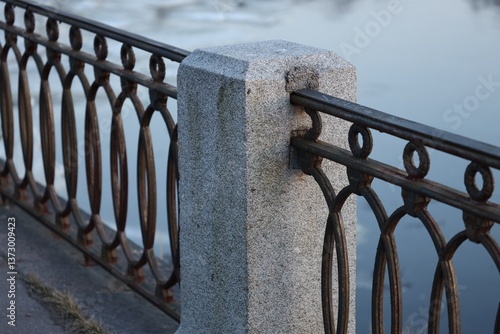 Fotografija A metal fence with a stone post situated beside a calm body of water
