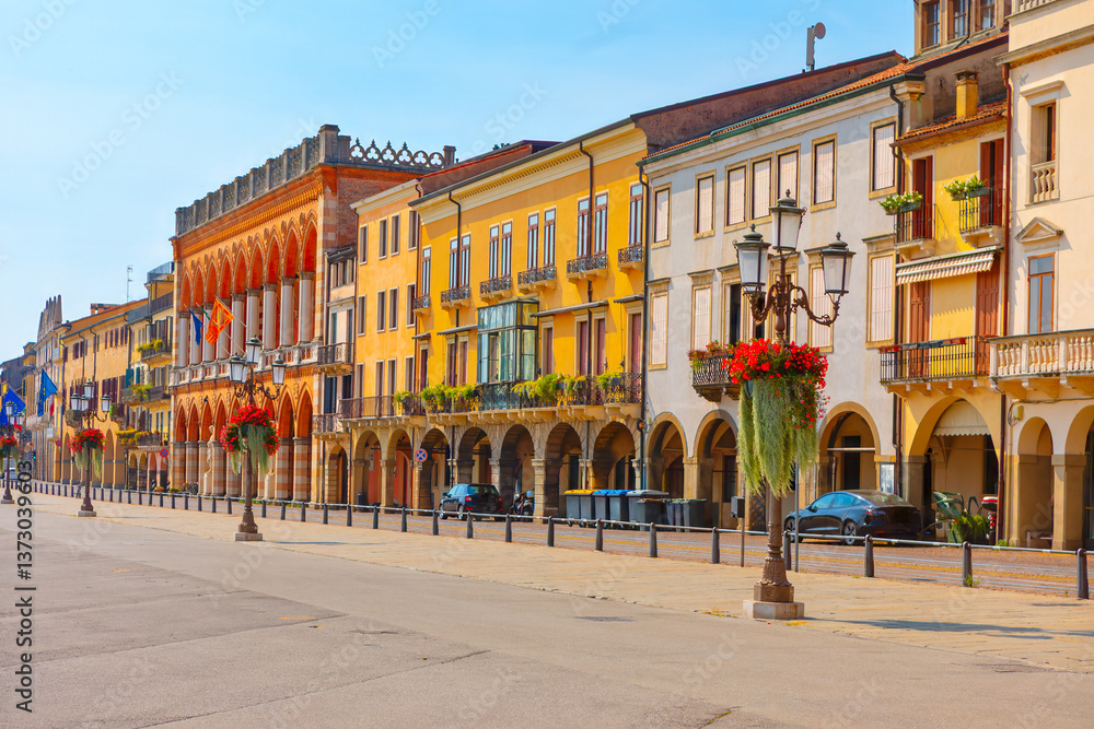 Charming street in Padua, Italy, lined with vibrant buildings featuring arches, balconies, and decorative plants. Hanging flower baskets on lampposts enhance the idyllic atmosphere of empty street
