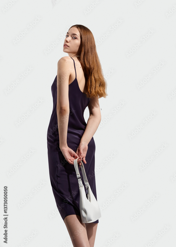Fototapeta premium A beautiful stylish woman with long well-groomed hair in a silk dress poses holding a fashionable white bag in her hands. The image of spring-summer 2025