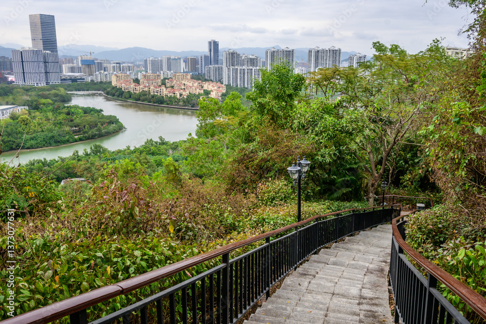 Fototapeta premium A view from a hilltop overlooking a cityscape with a river winding through it. Stone stairs with a metal railing lead down the hill towards a lush green landscape of Sanya, Hainan, China.