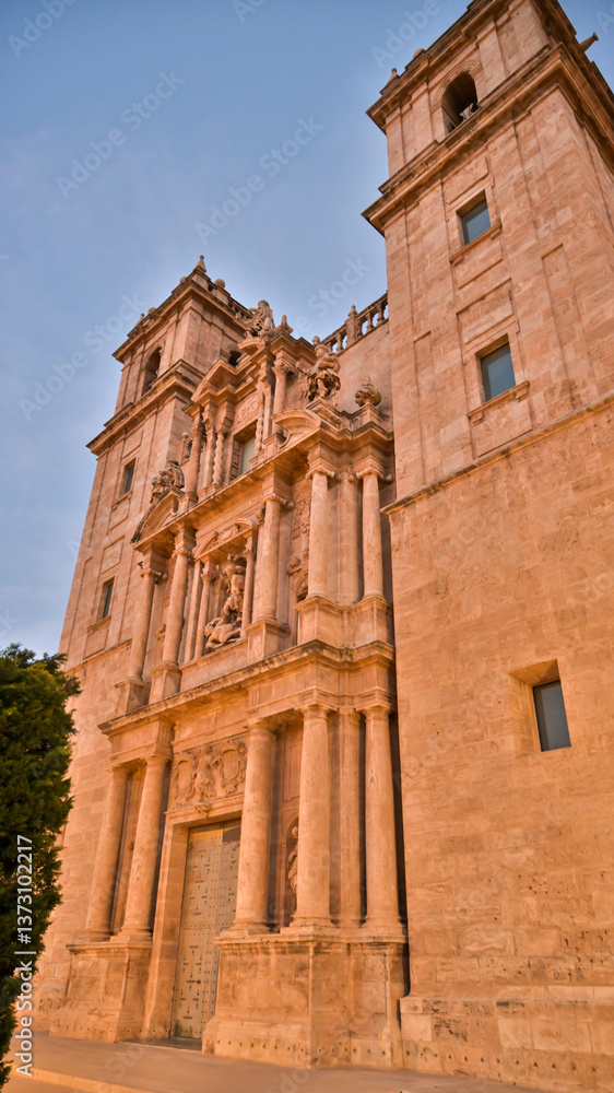 Fototapeta premium Iconic historic facade of Valencia Cathedral in warm evening light highlighting intricate stone carvings, symmetrical towers, and detailed architectural design.