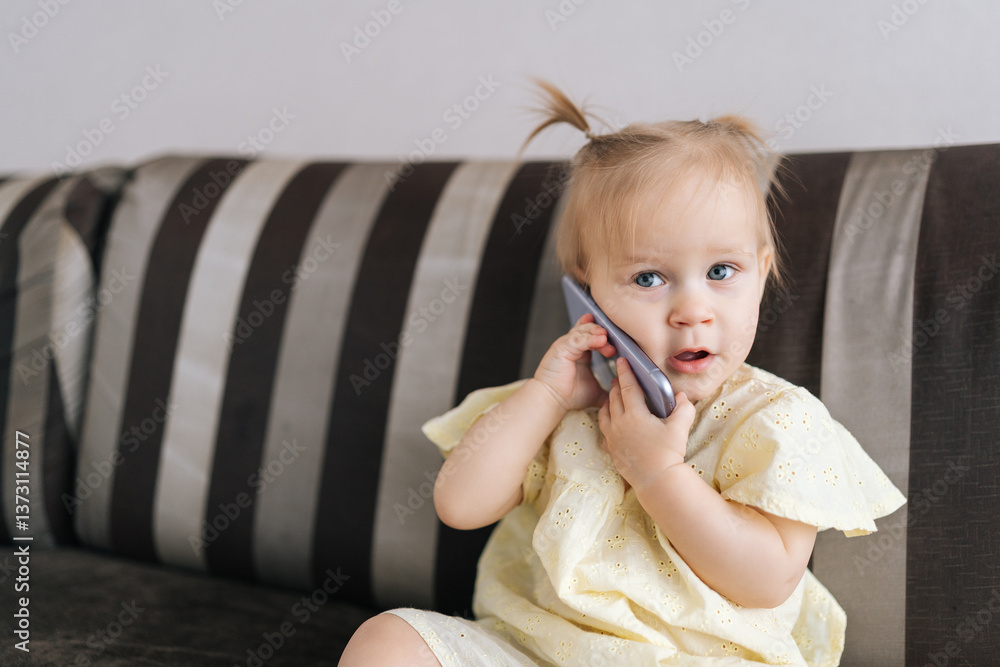 Cute toddler girl with small ponytail playfully talking using smartphone while sitting on sofa at home. Concept of imaginative play and early interaction with technology. Cute toddler girl with small ponytail playfully talking using smartphone while sitting on sofa at home. Concept of imaginative play and early interaction with technology.