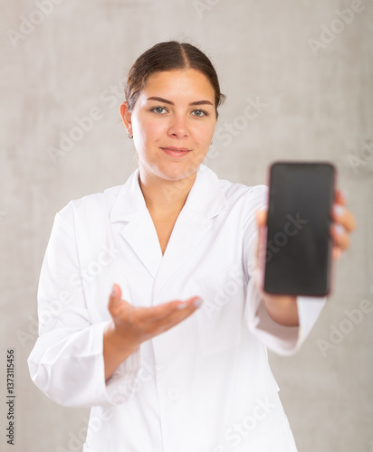 Принти на полотні Female doctor in white coat extending hand with phone and showing screen, selective focus on gadget