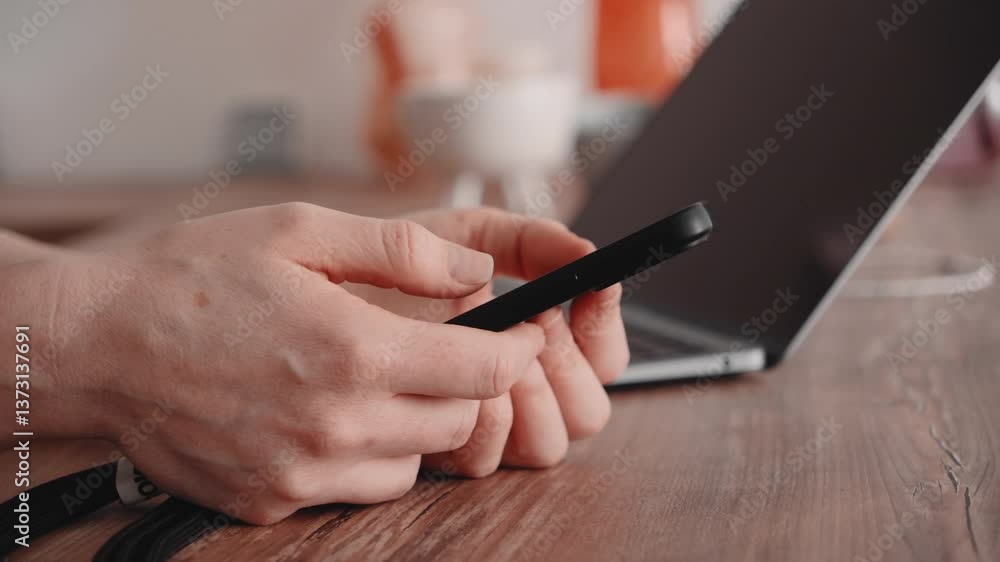 Female hands using mobile phone in kitchen. Slide left on smartphone against laptop background. Female hands using mobile phone in kitchen. Slide left on smartphone against laptop background.