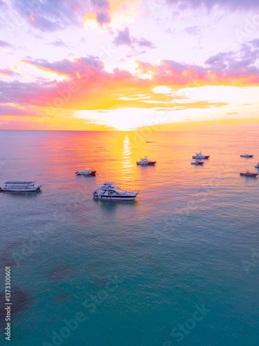 Aerial view of colorful sunrise over ocean in Punta Cana. Boats anchored in calm waters, surrounded by breathtaking scenery. Sky is painted with stunning hues, and gentle waves reflect morning light
