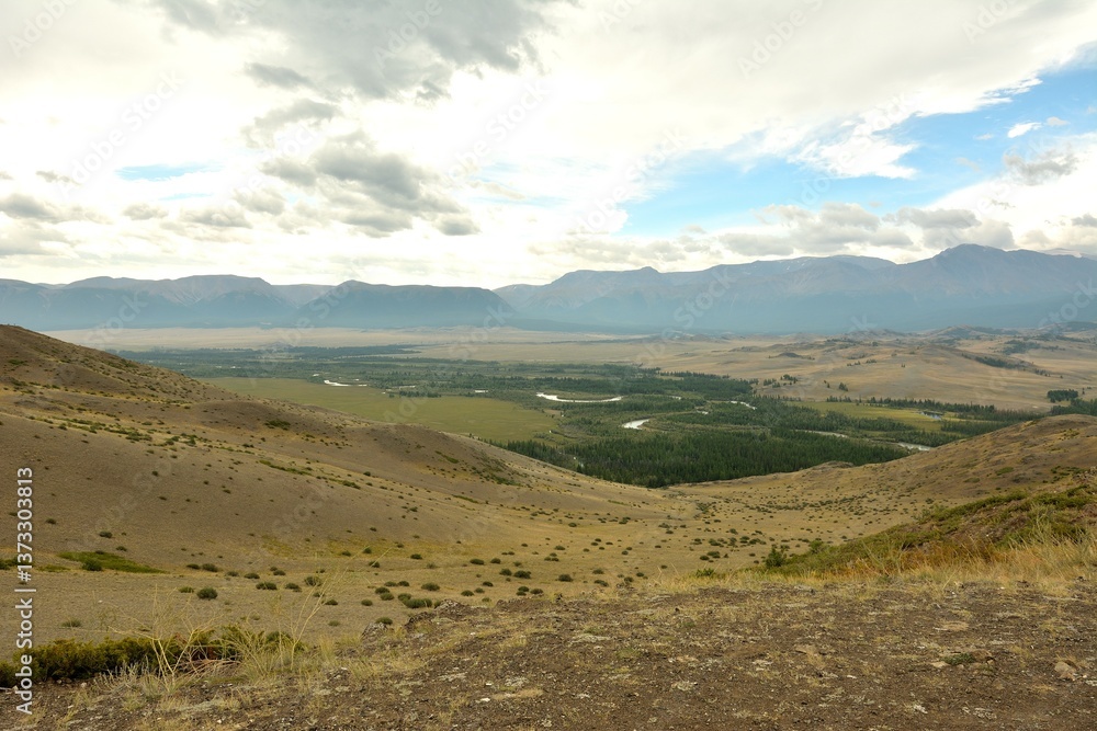 Fototapeta premium View of the endless steppe at the foot of snow-capped mountains through the junction of the foot of two hills on a cloudy summer day.