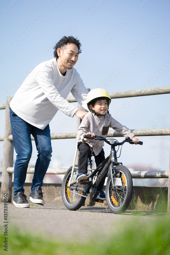 Obraz premium 青空の河川敷で川沿いで自転車の練習をする子どもと父親 がんばれ～