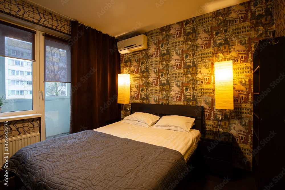 Apartment interior. Bedroom. Pillows and bedspread on the made bed. Wallpaper with images of European landmarks. Air conditioning and wall lamps on the wall. Window in the wall and door to the balcony