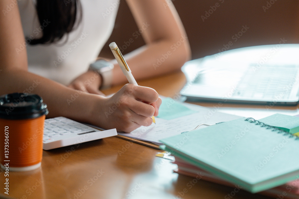 Businesswoman writing notes while working at desk with laptop and calculator Businesswoman writing notes while working at desk with laptop and calculator