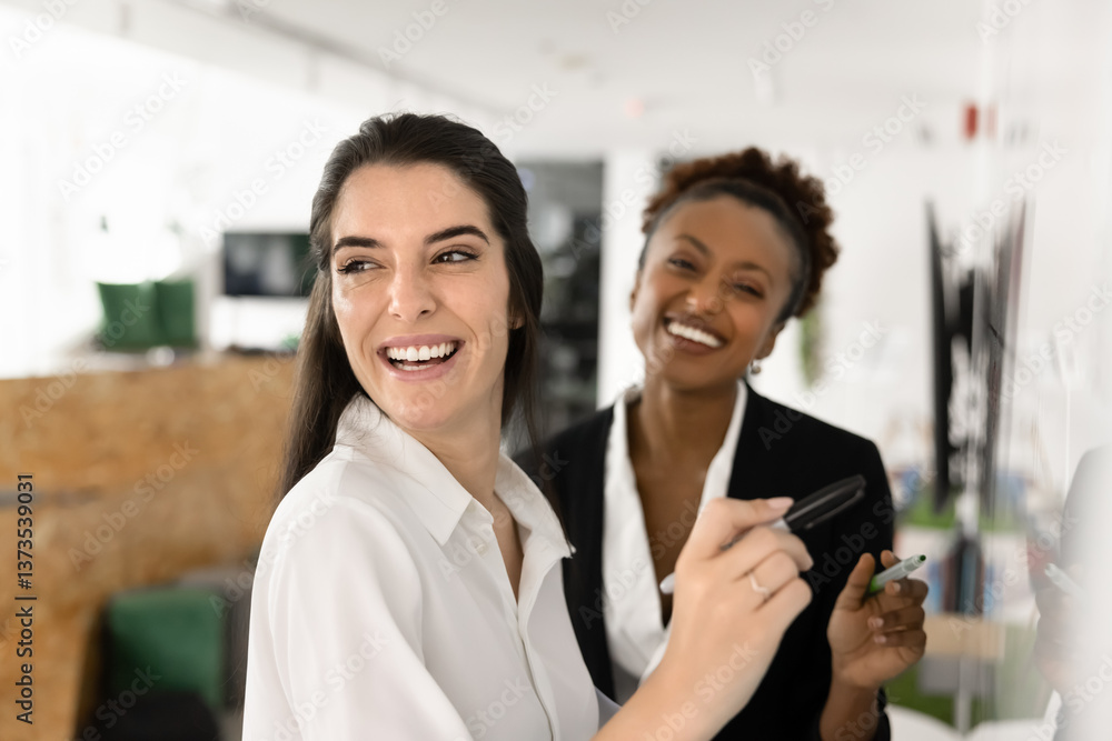 Obraz premium Cheerful diverse female coworkers writing teamwork plan on whiteboard, looking at camera and away, smiling, laughing. Office friends brainstorming, enjoying working together