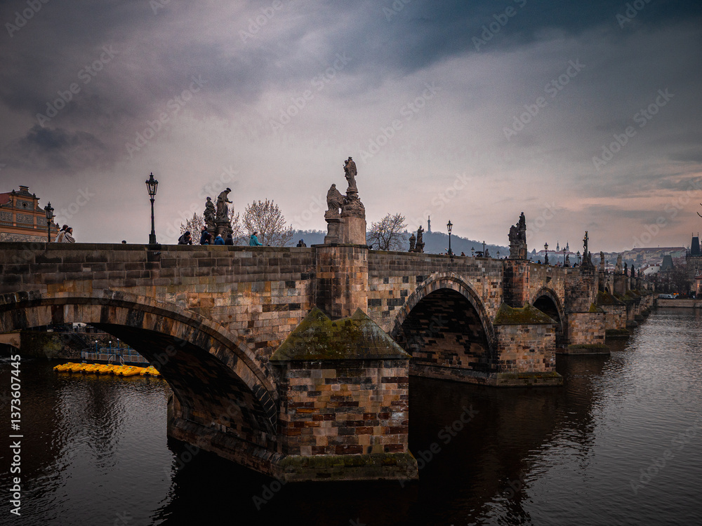 Obraz premium Charles Bridge in Prague during a moody evening with statues and arches reflected in the Vltava River, capturing a historic and romantic atmosphere.