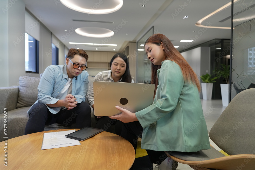 A 29-year-old Asian woman in a suit holds a laptop explaining the terms and conditions of life insurance to male and female customers in an office lounge.