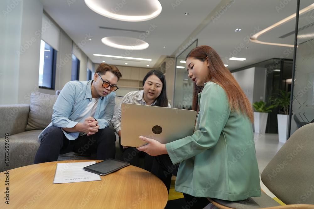 A 29-year-old Asian woman in a suit holds a laptop explaining the terms and conditions of life insurance to male and female customers in an office lounge.