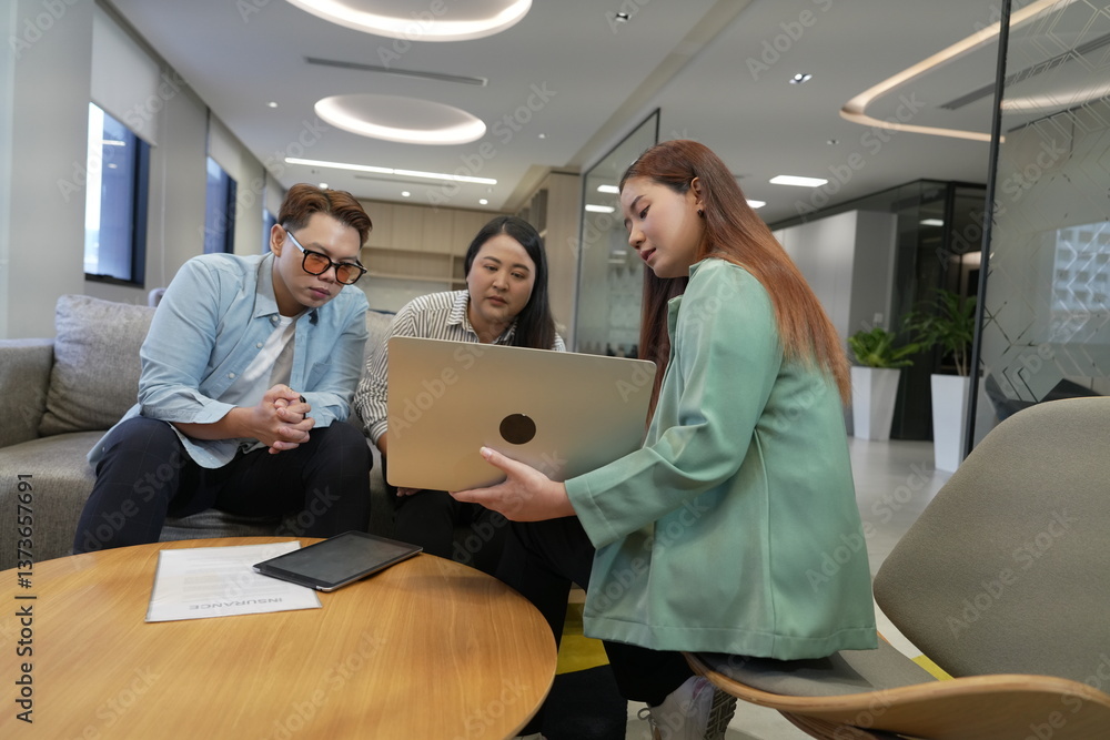 A 29-year-old Asian woman in a suit holds a laptop explaining the terms and conditions of life insurance to male and female customers in an office lounge.