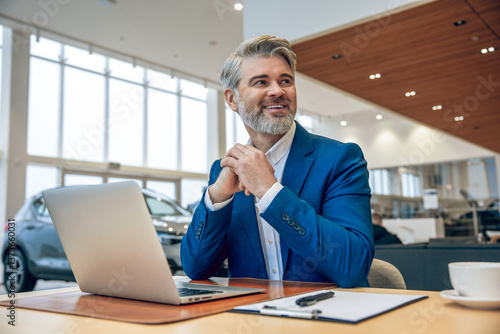 Bearded salesman in suit working on his laptop at new cars showroom